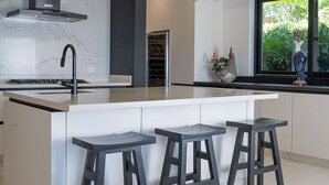 Una cocina moderna con una isla central, taburetes de bar, lámparas colgantes y un amplio ventanal con vistas a la vegetación.
