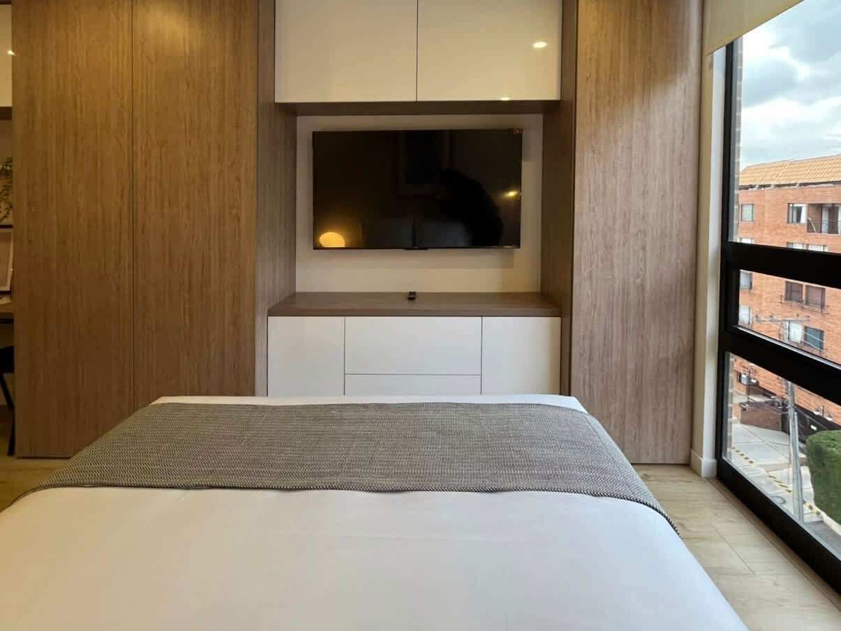 A modern bedroom with a large bed, a built-in TV, and a view of the city through large windows.