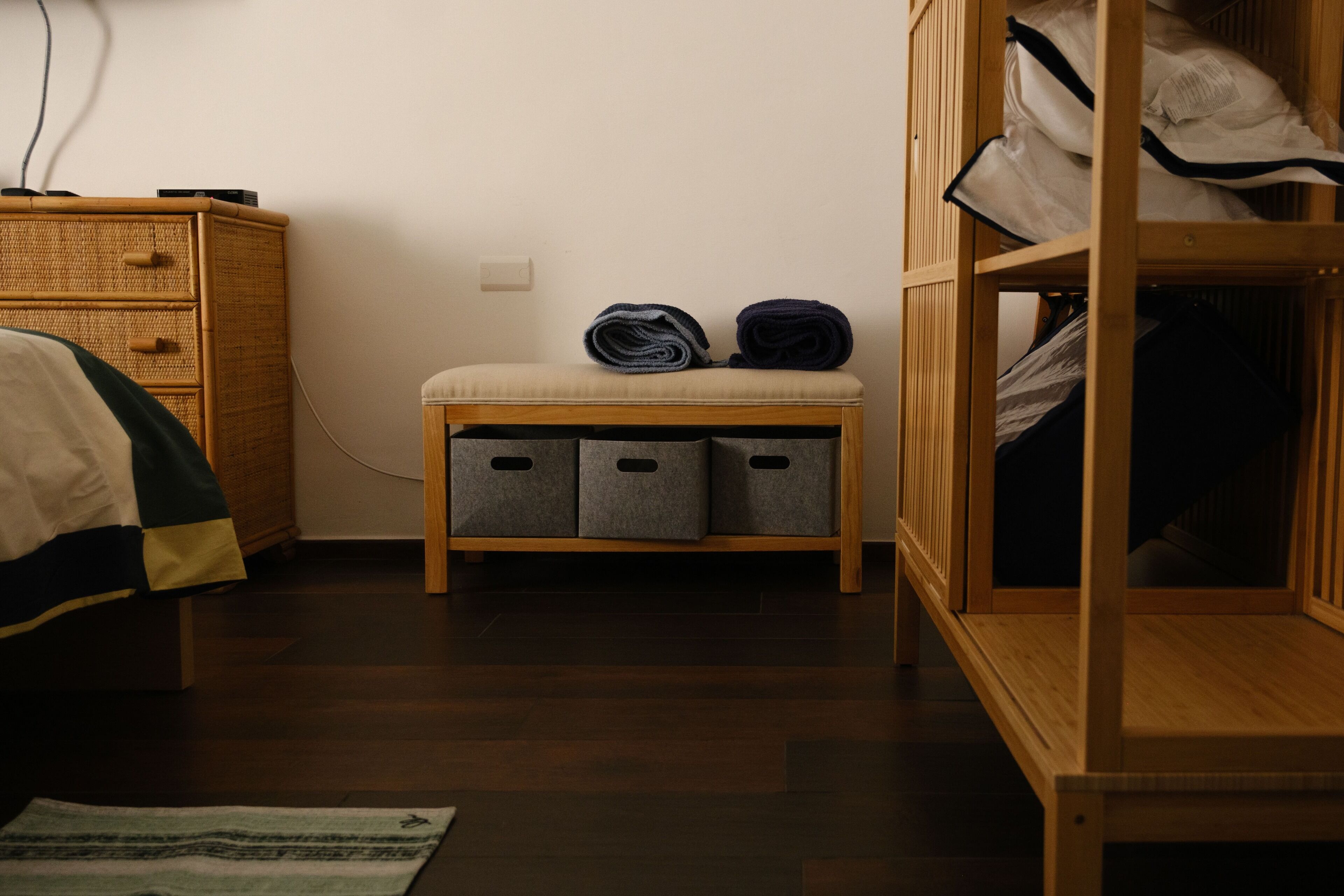 A bedroom with a wooden wardrobe, a bedside table with storage bins, a bed with a striped blanket, and a wall socket.