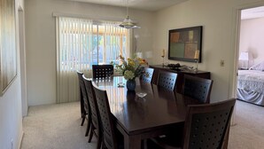 A hotel room with a dining area featuring a wooden table, chairs, and a vase of flowers. There is a large window with blinds, a painting on the wall, and a bed with a quilted cover. - Charming 3-bedroom house with AC in sunny Scottsdale (Scottsdale)