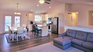 A spacious living area with a grey sofa, a dining table with chairs, a kitchen with modern appliances, and a ceiling fan.