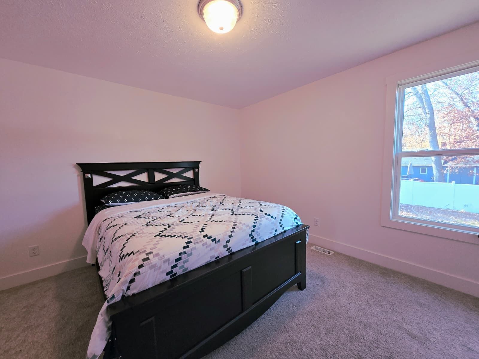 A bedroom with a black bed frame, a patterned bedspread, and a window with a view of trees and a fence.