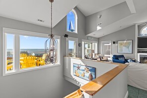 A room with a view of the ocean, a large window, a wooden bench, and a framed picture of sea shells. - Ocean Views! New 4bd Home, very close to Beach! (Oak Island)