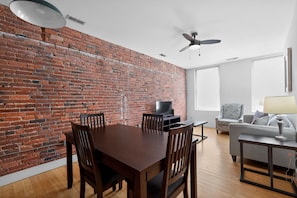 A room with a brick wall, a dining table with chairs, a sofa, a TV, and a ceiling fan.