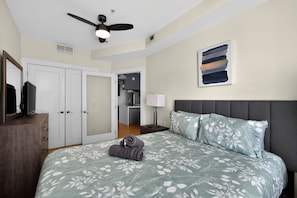 A bedroom with a bed, a ceiling fan, a TV, a wardrobe, a painting, and a kitchen area.