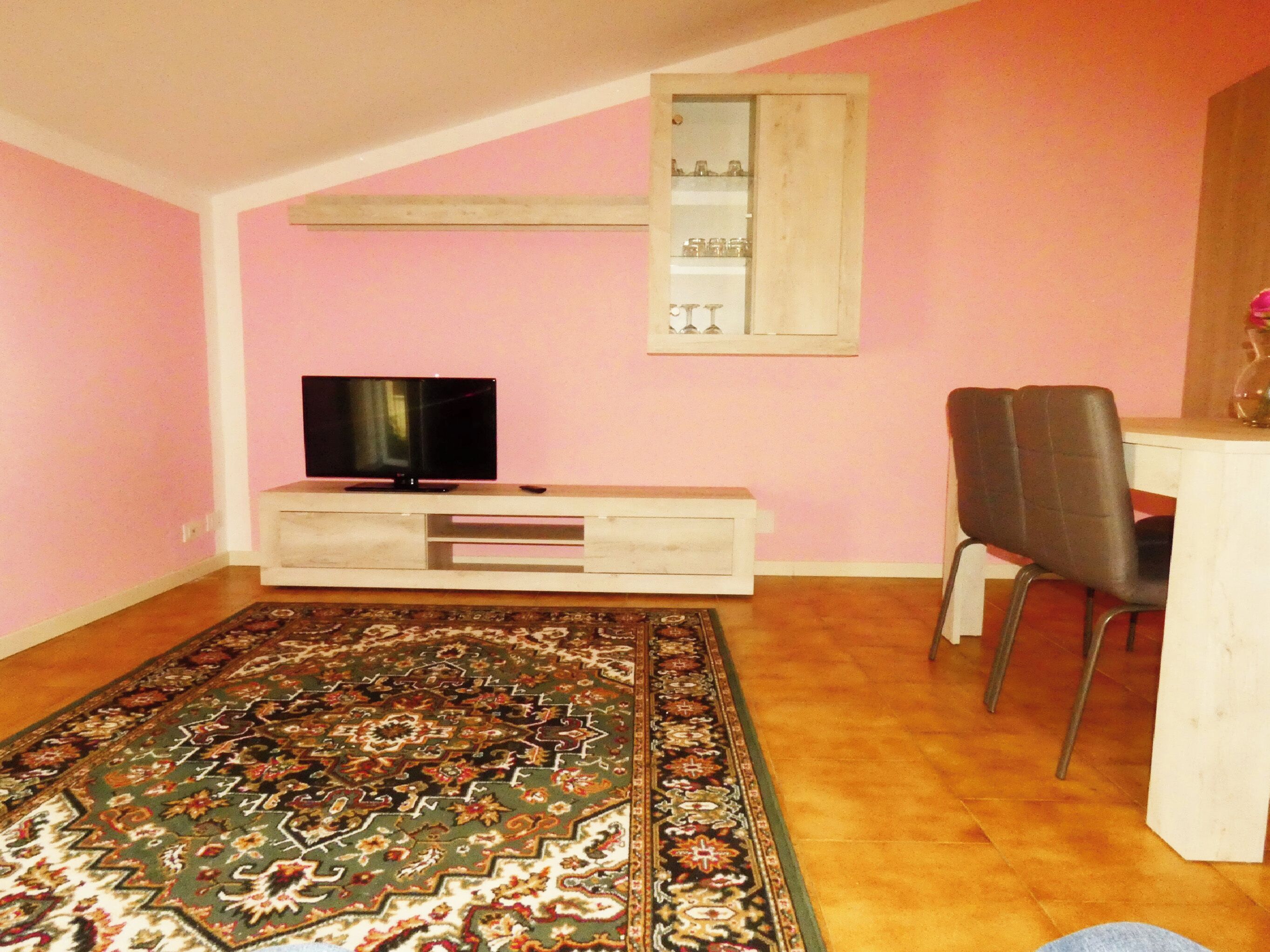 Een kamer met een televisie op een witte kast, een zithoek met een tafel en stoelen, en een kamerbreed tapijt op de vloer.