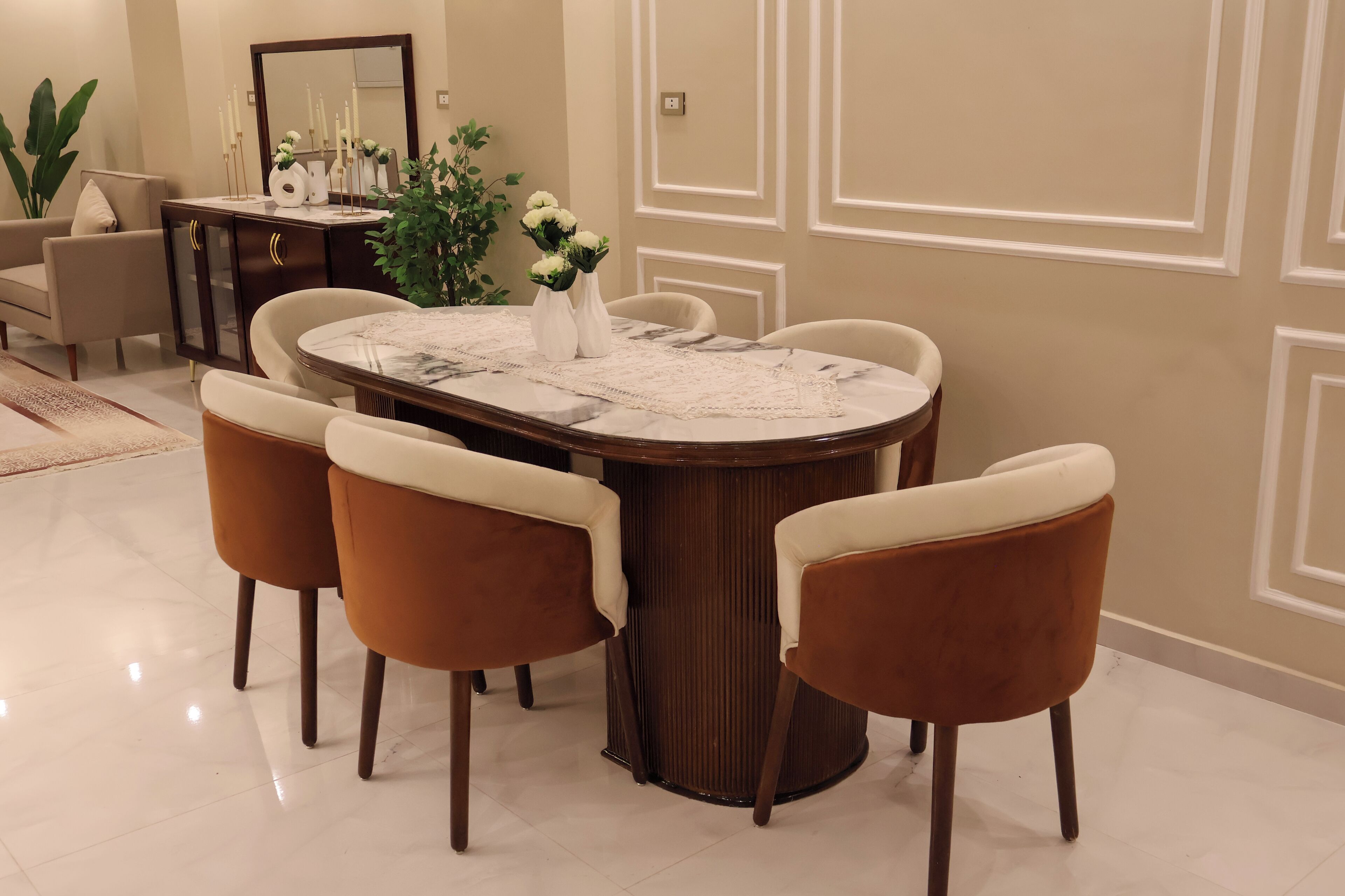 Un comedor con una mesa redonda, cuatro sillas, un florero con flores y un aparador con objetos decorativos.