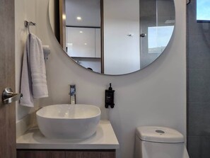 Une salle de bain moderne avec un grand lavabo ovale, un miroir rond, un porte-serviettes, un sèche-mains et des toilettes.