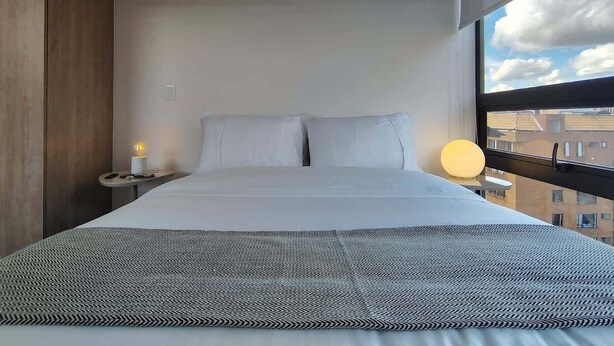 Ein modernes Hotelzimmer mit einem großen Bett, einem Nachttisch, einer Nachttischlampe und einem Fenster mit Blick auf die Stadt.