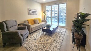 Un salon avec un canapé gris, une table basse, un tapis à motifs et une grande fenêtre donnant sur un immeuble.