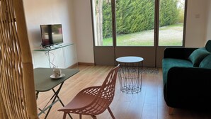 En moderne stue med en grøn sofa, et lille rundt bord, et fjernsyn på et glas skab og et stort vindue med udsigt til træer og græs.