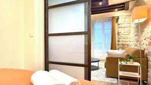 A hotel room with a bed, a wooden floor, a stone wall, a small table with a chair, and a view of the outside. - Paris Loft for 4 by Châtelet & Montorgueil & Metro (PARIS)