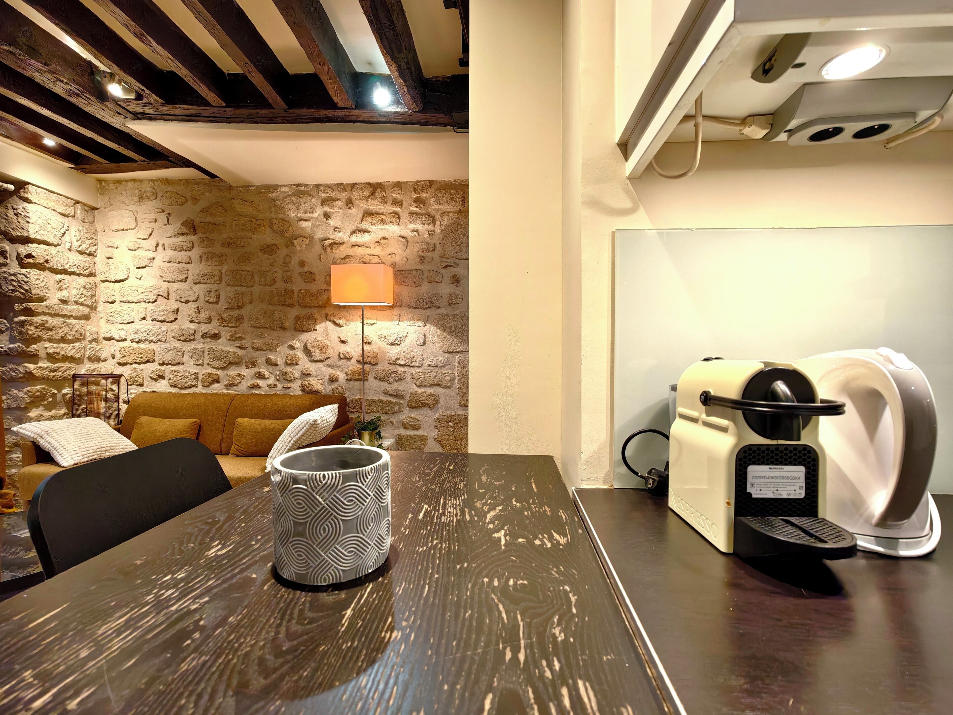 A cozy room with a stone wall, a wooden ceiling, a sofa, a coffee machine, and a patterned mug on a table.