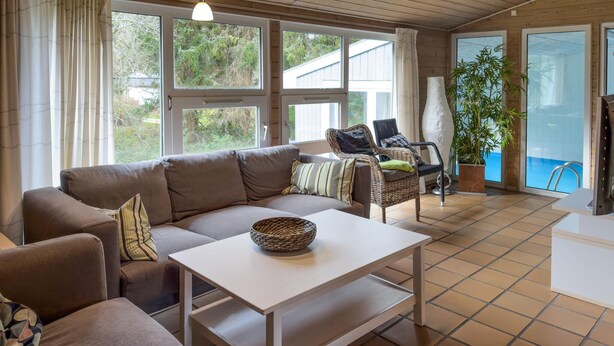 A cozy living room with a sofa, a coffee table, and a wicker chair. There is a large window with curtains, a potted plant, and a view of a pool. - Stunning home in Knebel (Knebel)