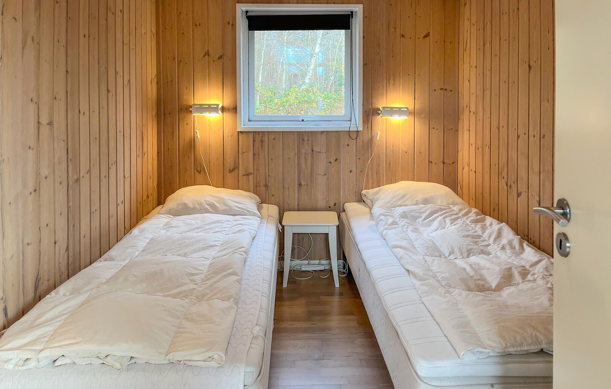 A small, cozy room with wooden walls and a window, featuring two single beds and a small table.