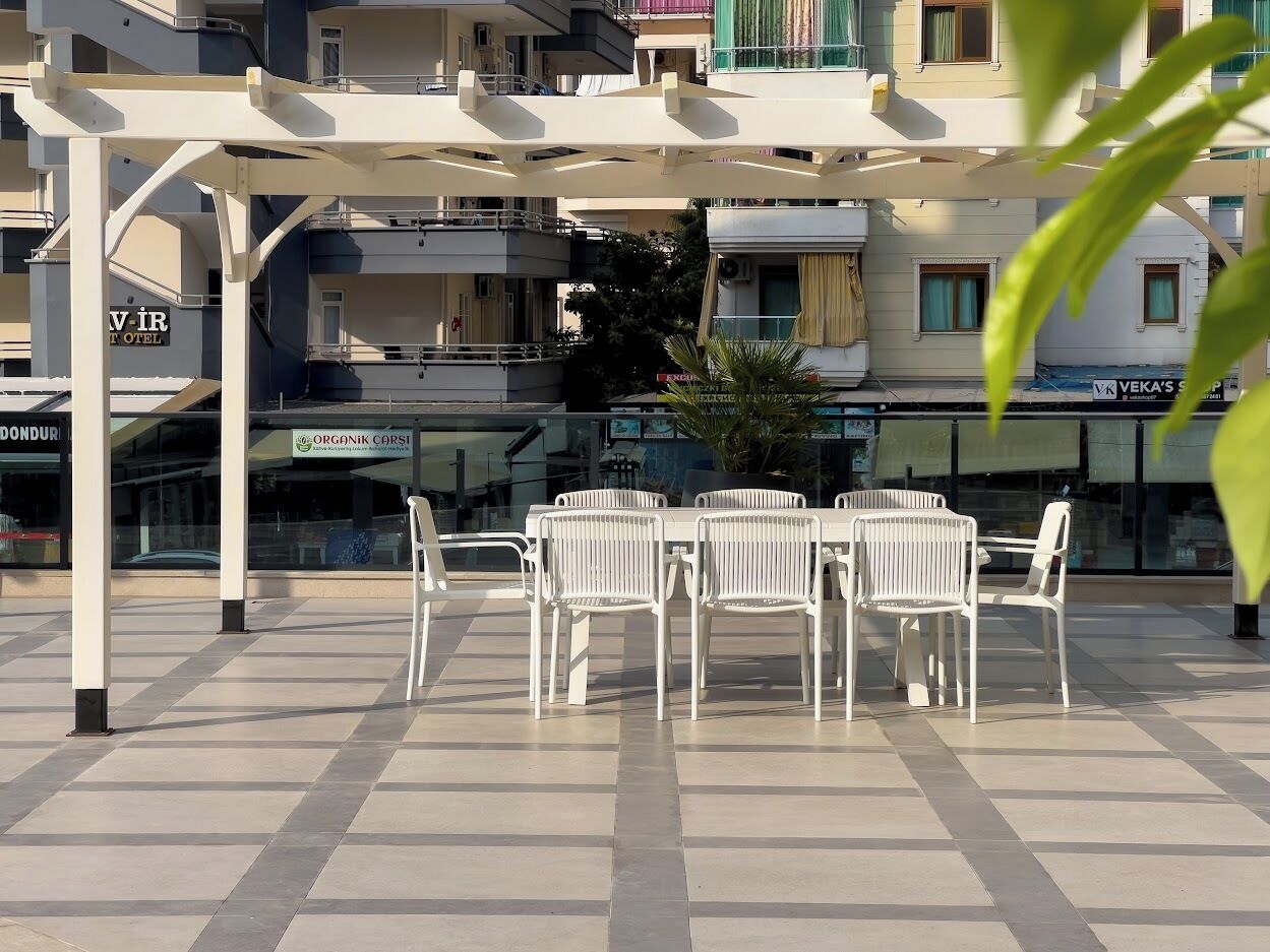 Eine Terrasse mit weißen Metallstühlen und Tischen, einer Pergola darüber und Blick auf ein Gebäude mit Balkonen und Geschäften im Hintergrund.