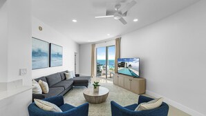 Ein modernes Wohnzimmer mit einem grauen Ecksofa, zwei blauen Sessel, einem runden Couchtisch und einem Flachbildfernseher auf einer Holzkommode.