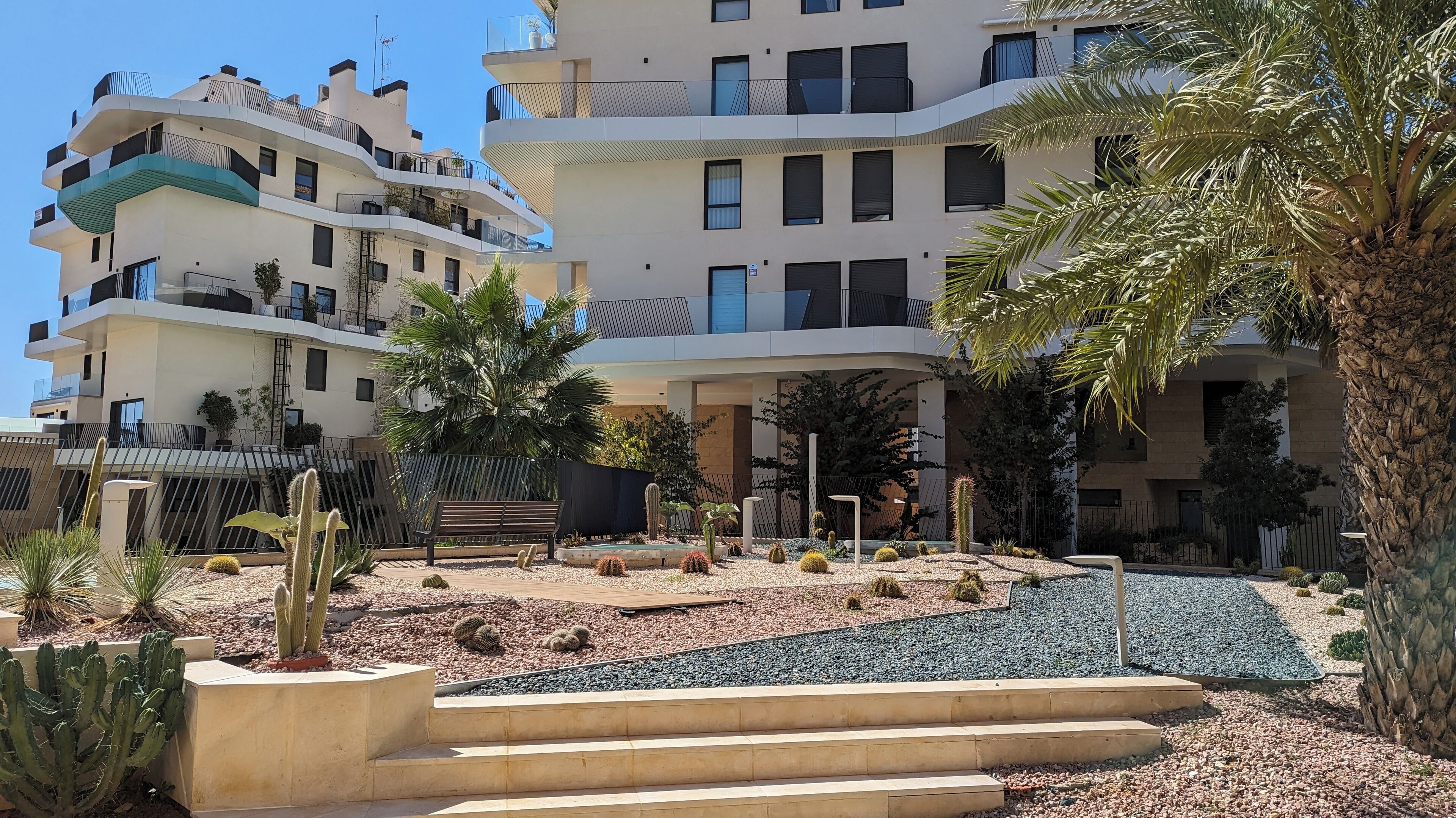 Un moderno edificio residencial de varios pisos con balcones, un jardín ajardinado con cactus y palmeras, y un banco de madera.