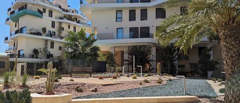 Un moderno edificio residencial de varios pisos con balcones, un jardín ajardinado con cactus y palmeras, y un banco de madera.