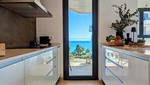 A modern kitchen with a view of the sea, a dining area with a table and chairs, and a balcony with plants. - Aster Apartments (Villajoyosa)