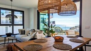 A modern living room with a large wooden dining table, woven placemats, and a view of the sea. - Aster Apartments (Villajoyosa)