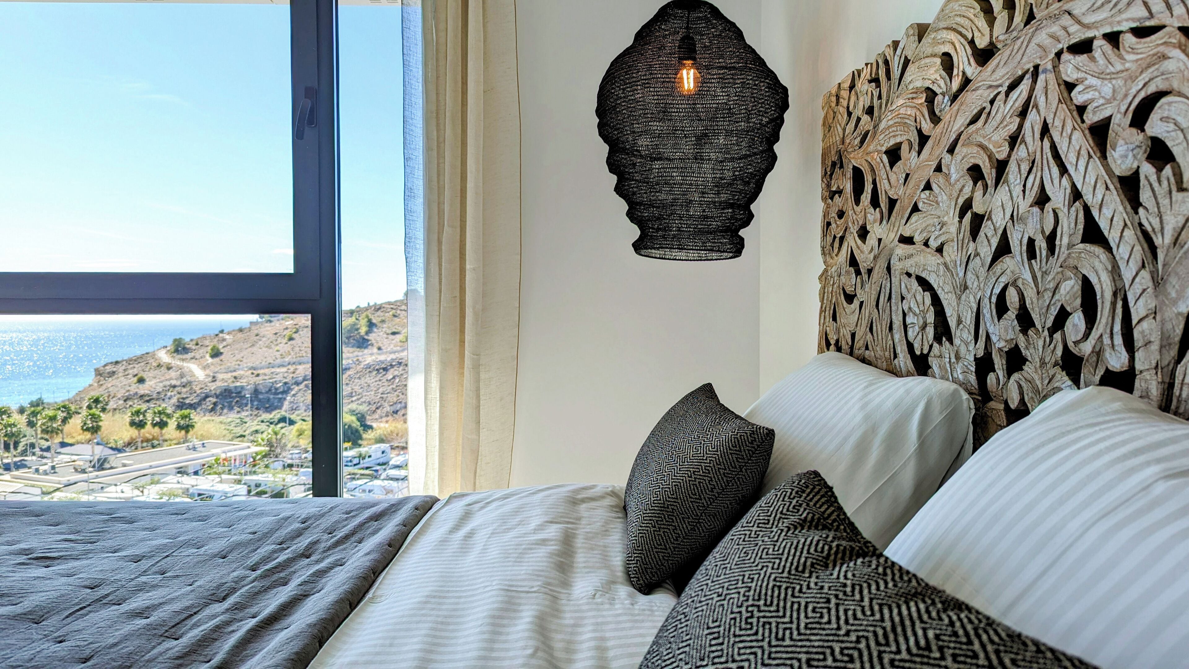 Una cama con almohadas estampadas, un cabecero con textura y una lámpara colgante.