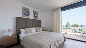 A bedroom with a large bed, two bedside tables with lamps, and a view of the sea through a large window. - Aster Apartments (Villajoyosa)