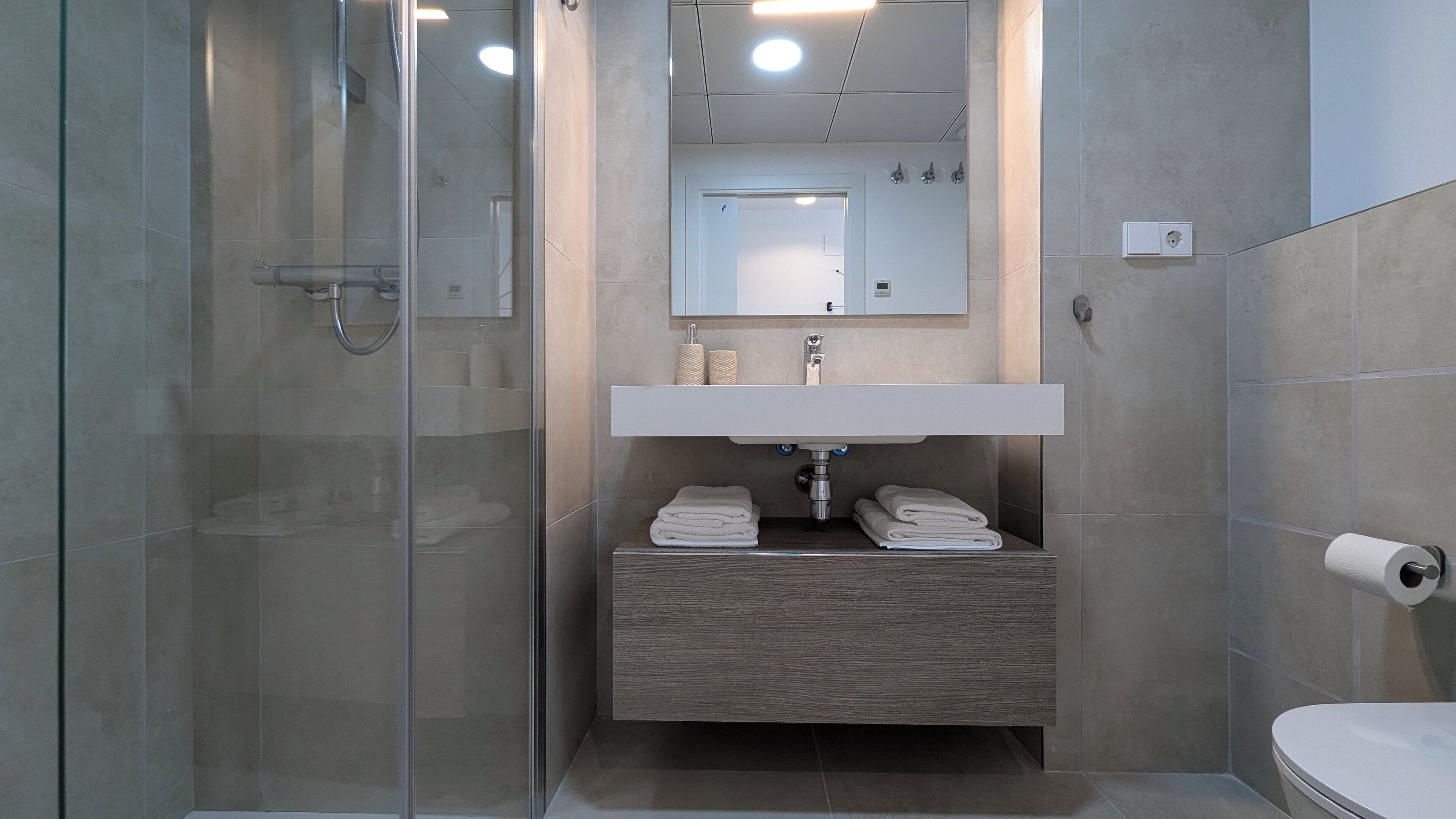Un baño moderno con cabina de ducha de vidrio, tocador de madera, lavamanos blanco y retrete.