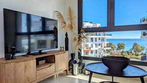 A modern living room with a large flat-screen TV on a wooden console, a small table with a black chair, and tall decorative plants. A large window offers a view of a coastal building and the sea. - Aster Apartments (Villajoyosa)