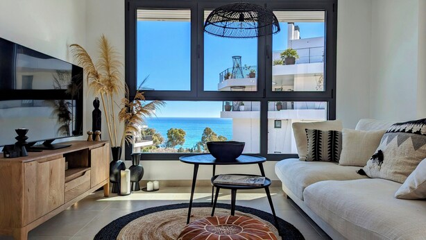 A modern living room with a large window overlooking the sea, a flat-screen TV, a wooden cabinet, and a comfortable sofa. - Aster Apartments (Villajoyosa)