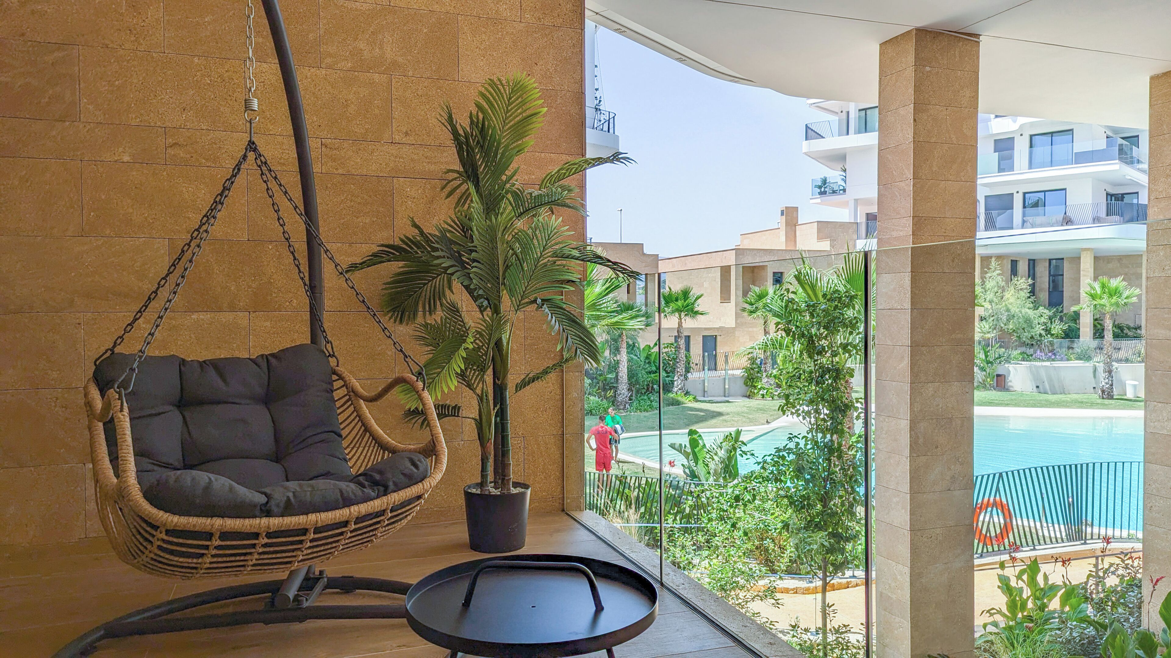 Un balcon avec un fauteuil suspendu en osier, un pot de fleurs et une vue sur une piscine et des immeubles.