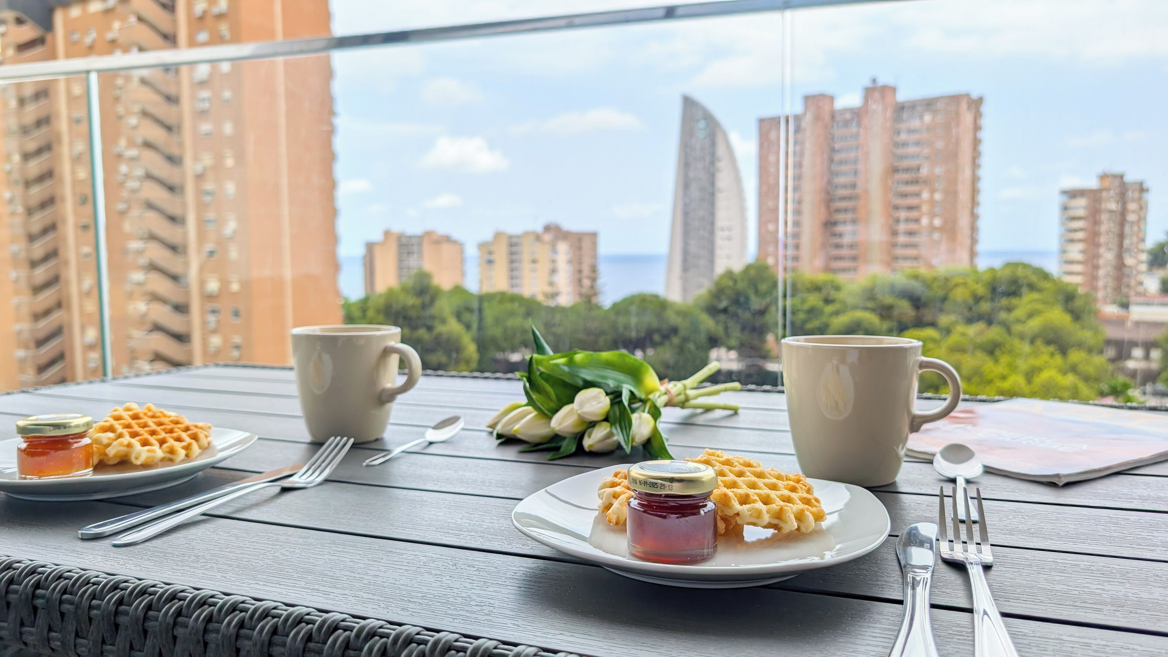 Mesa na varanda com waffles, geleia e xícaras de café, com vista para um horizonte urbano com edifícios altos.