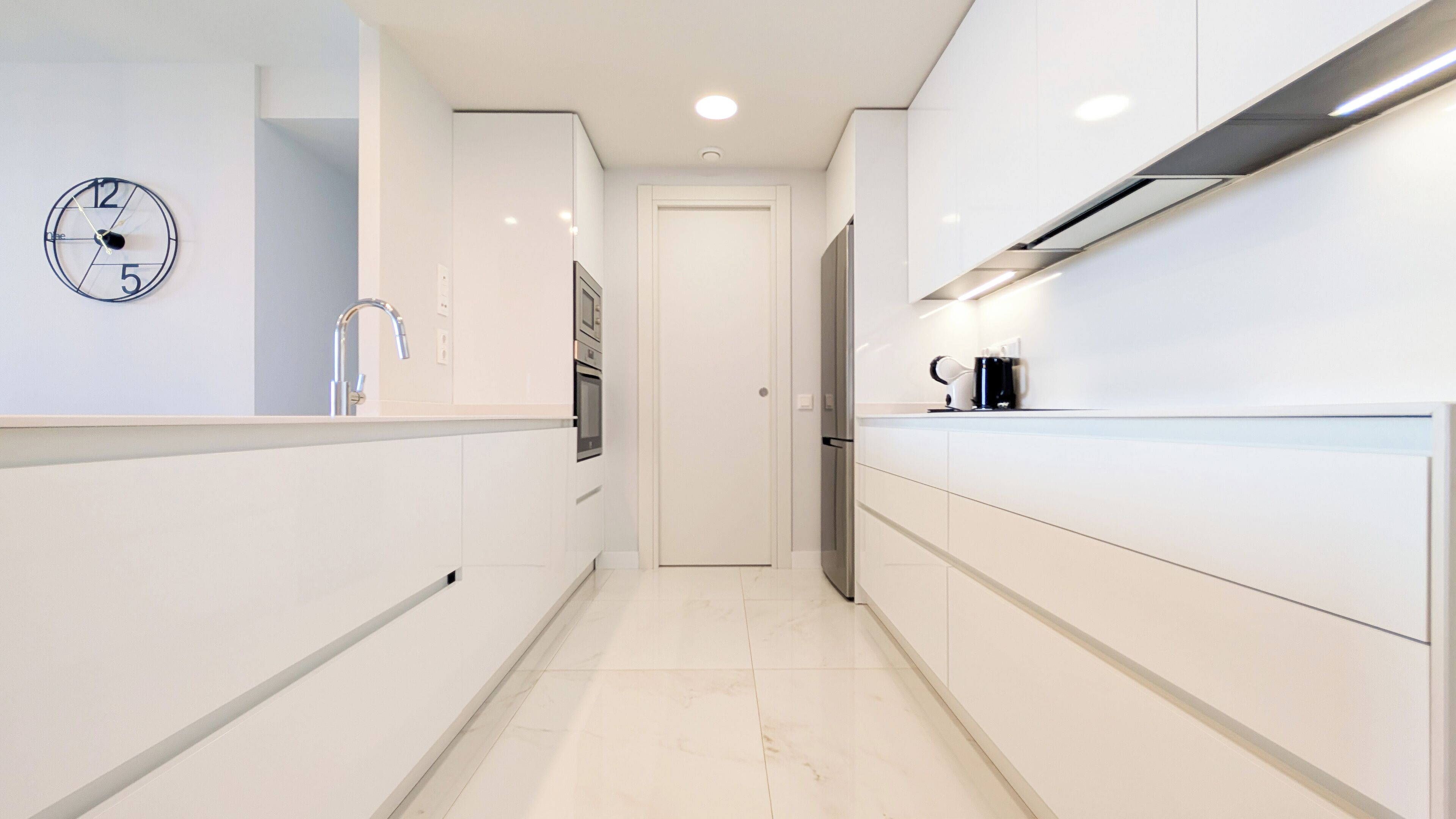 A modern kitchen with white cabinetry, a stainless steel refrigerator, a coffee maker, and a wall clock.