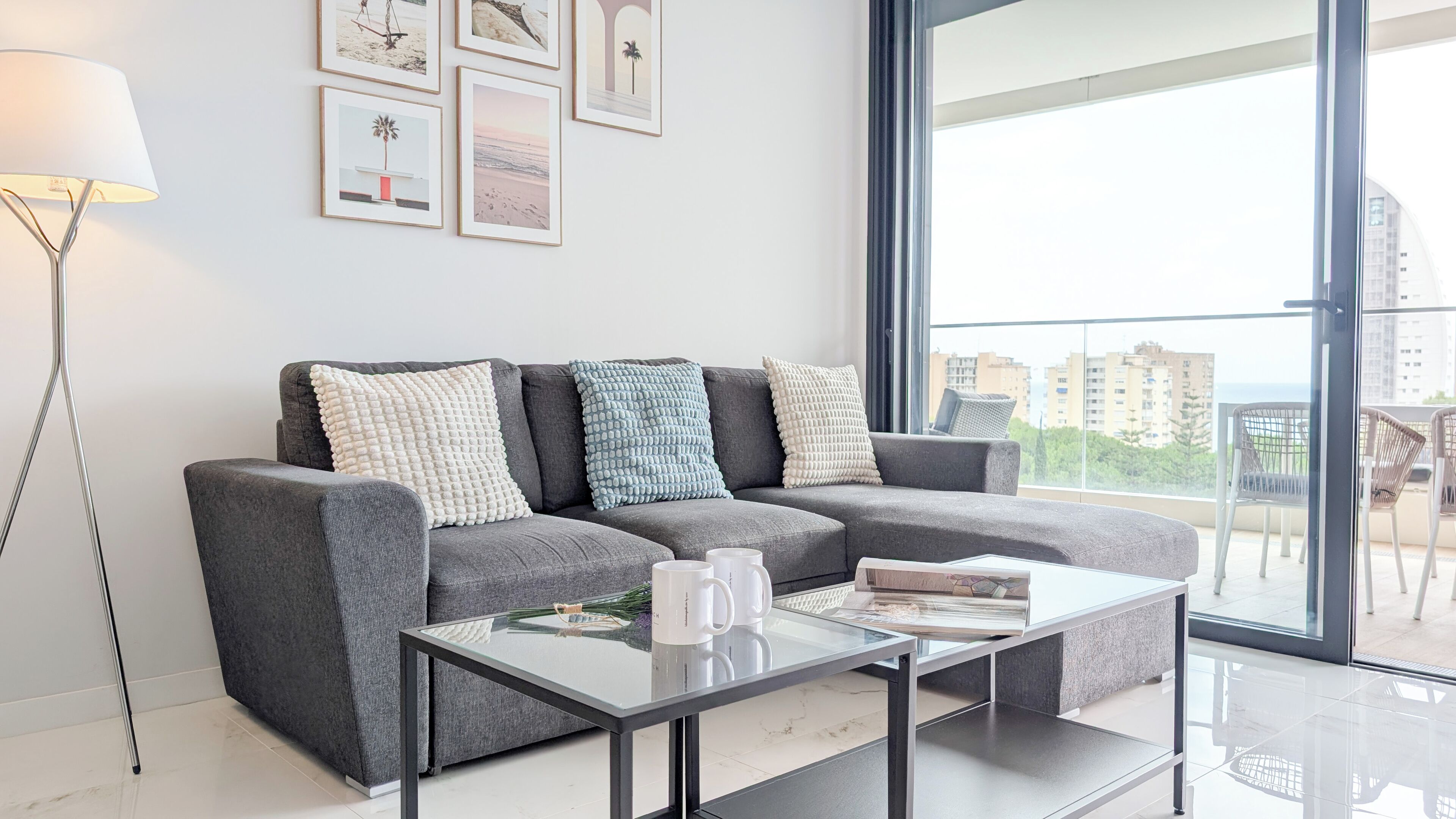 Un salon moderne avec un canapé gris, une table basse en verre et un lampadaire. Des tableaux encadrés ornent le mur, et un balcon offre une vue sur les bâtiments.
