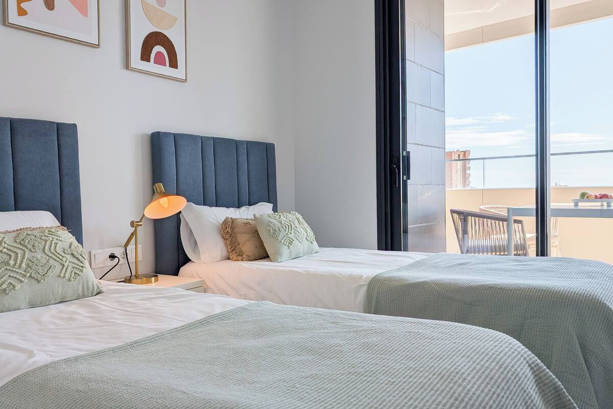 Quarto de hotel com duas camas, uma janela grande e varanda com mesa e cadeiras.