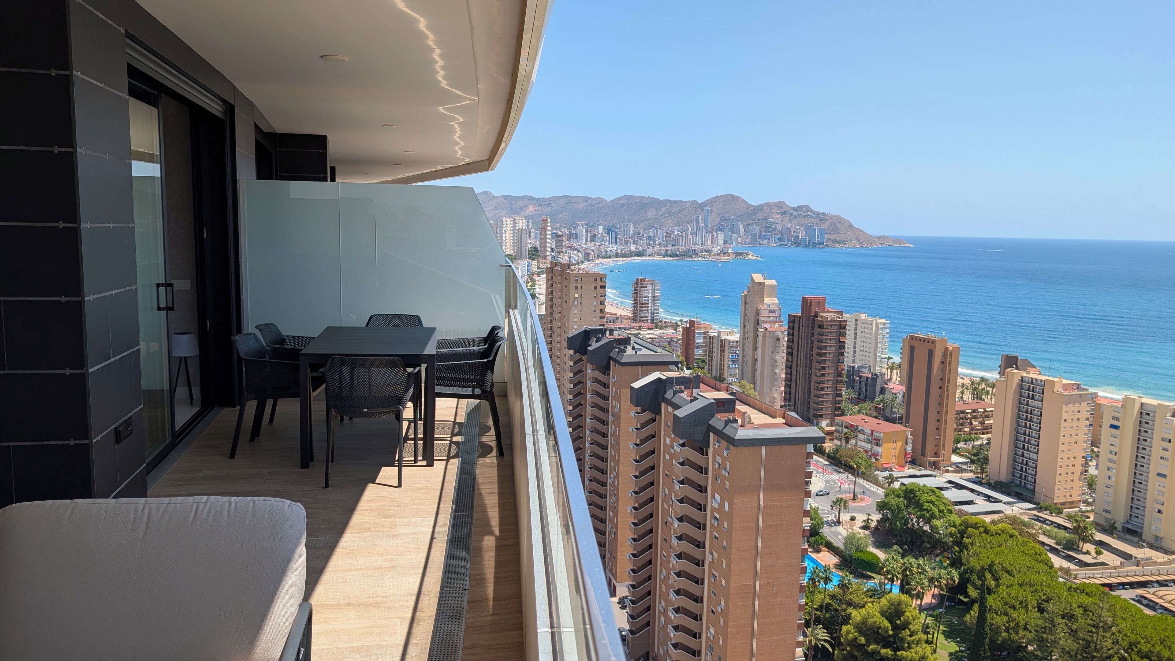 A balcony with a table and chairs, overlooking a coastal cityscape with high-rise buildings and a beach.