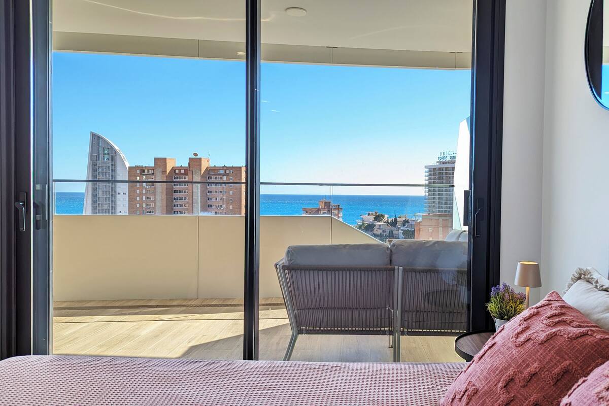 Uma varanda com vista para o mar e edifícios, uma cama com cabeceira e uma cadeira.