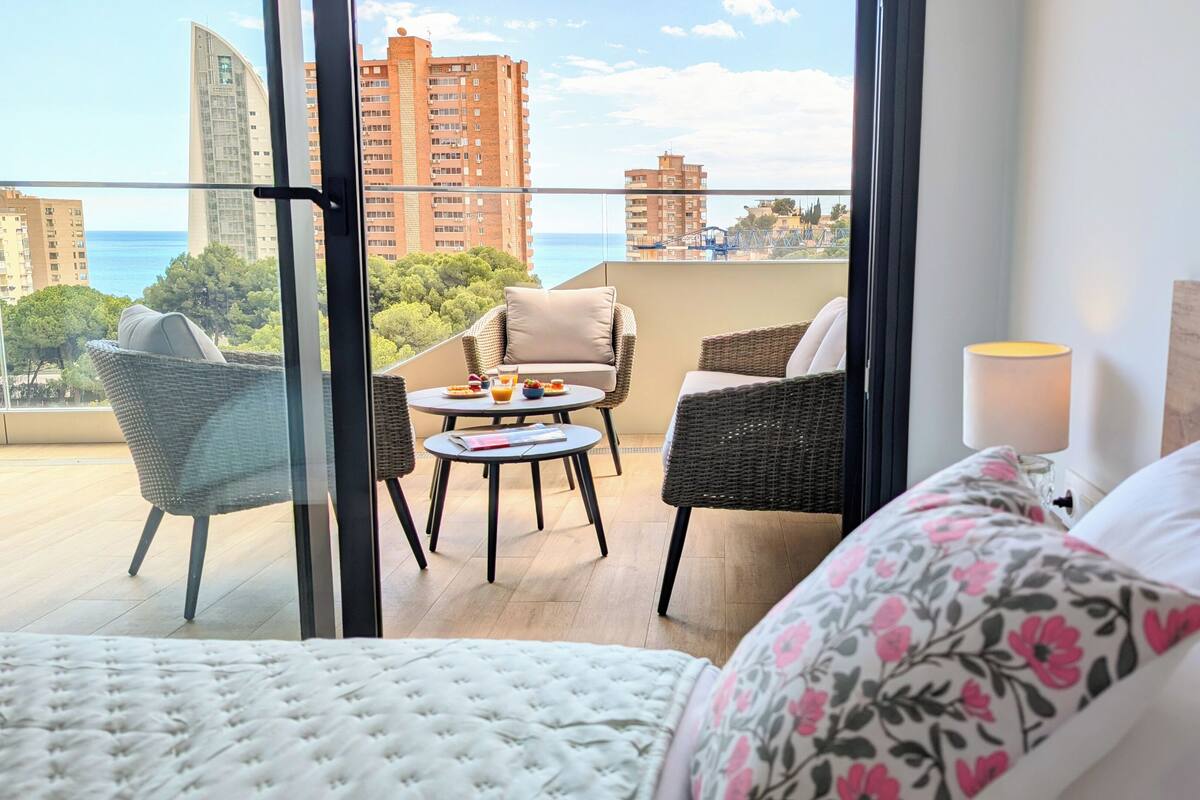 Quarto de hotel com varanda, móveis de vime, uma mesa pequena com café da manhã e vista para o mar e edifícios.
