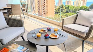 A balcony with wicker furniture, a table with breakfast items, and a view of high-rise buildings. - Benidorm Beach Apartments (Benidorm)