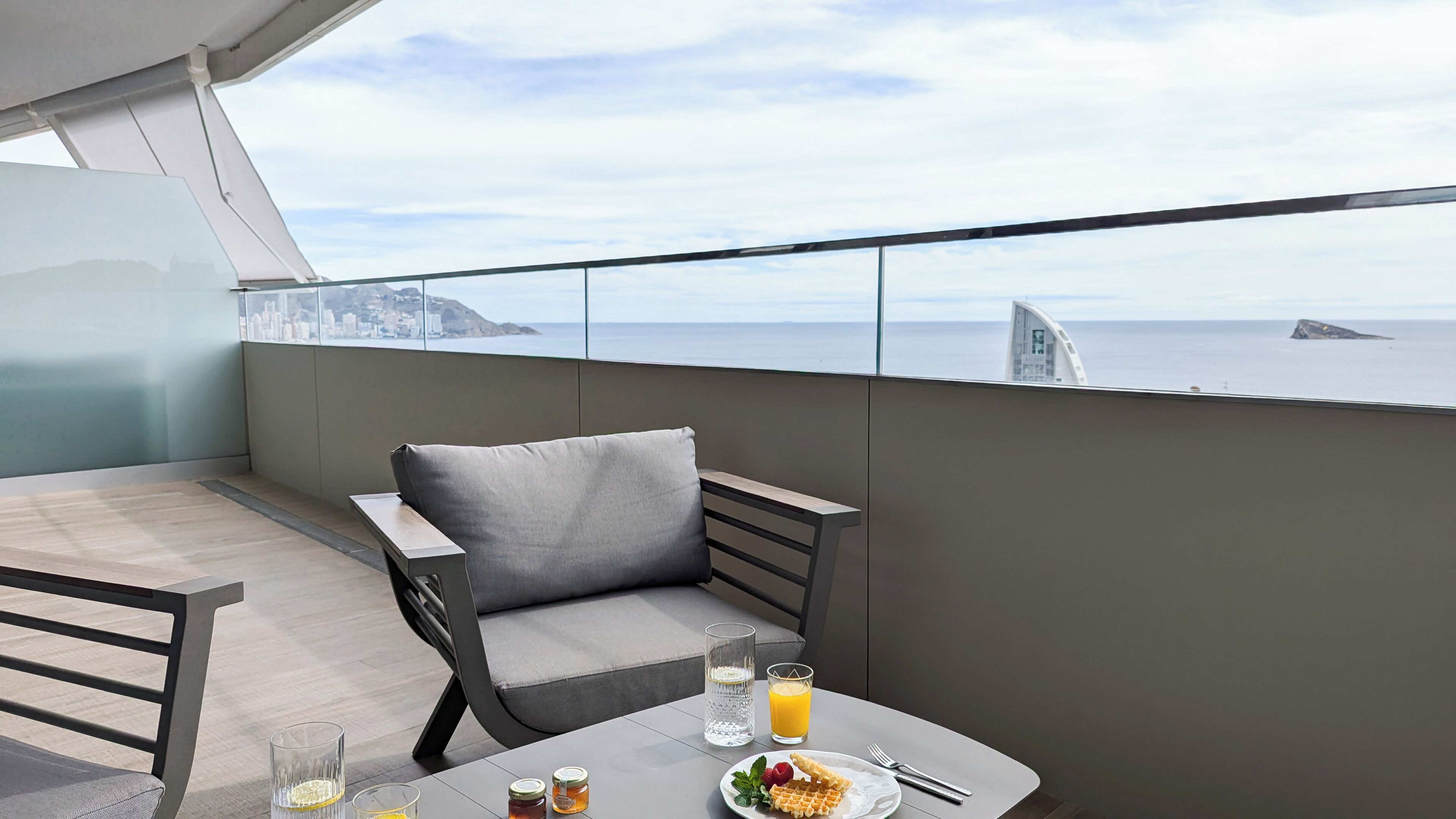 A balcony with a table set for two, featuring a glass of water, a glass of orange juice, a plate with waffles, and a jar of honey.