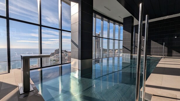 A modern indoor swimming pool with large windows offering a view of the sea and cityscape. - Benidorm Beach Apartments (Benidorm)