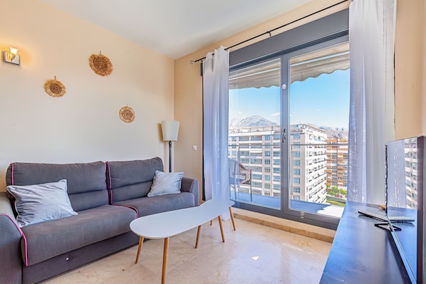 A modern living room with a sofa, a small table, a TV, and a large window with a view of buildings. - Cala Alta Apartments (Villajoyosa)