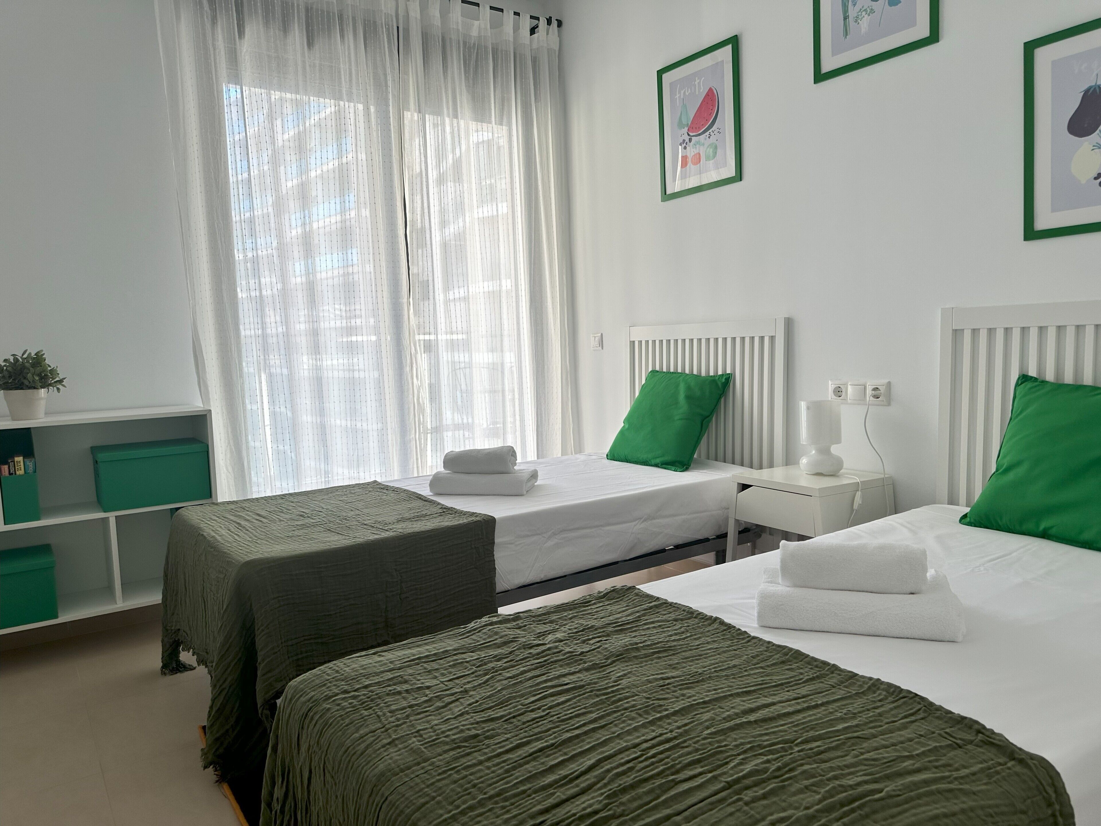 Une chambre d’hôtel avec deux lits, une table de chevet avec une lampe, une fenêtre avec des rideaux légers et un mur orné de tableaux.