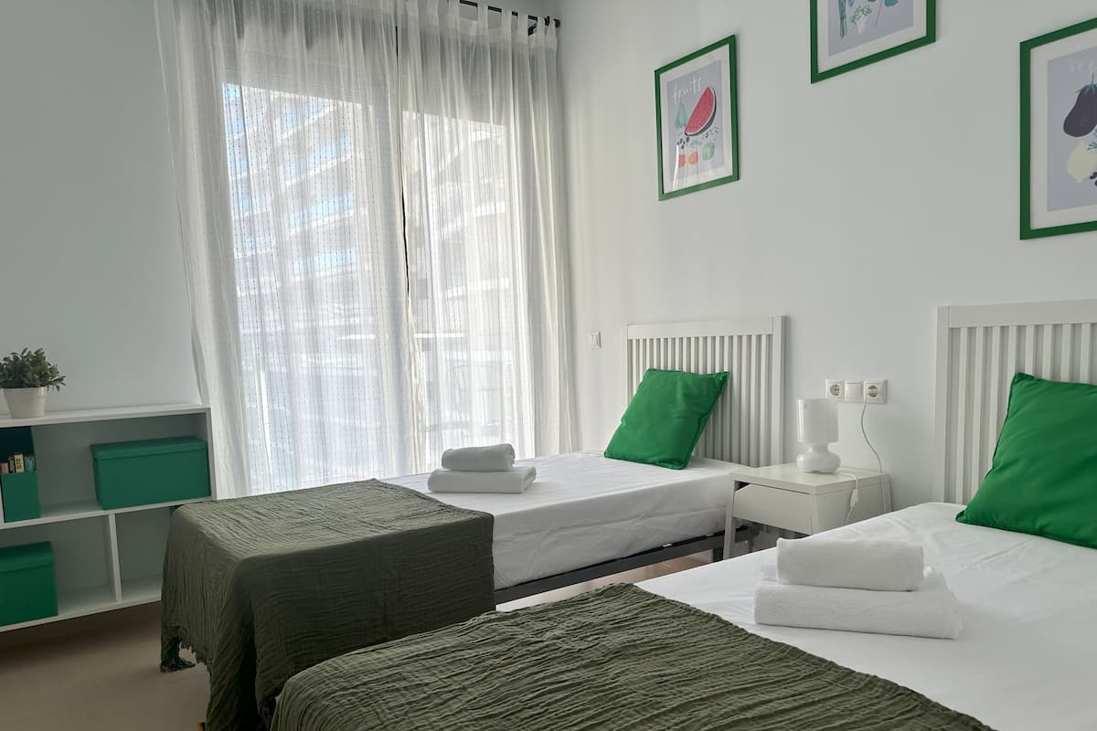 Une chambre d’hôtel avec deux lits, une table de chevet avec une lampe, une fenêtre avec des rideaux légers et un mur orné de tableaux.