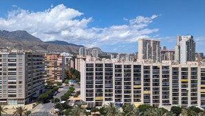 Um edifício residencial alto com várias varandas, uma rua em frente e montanhas ao fundo.