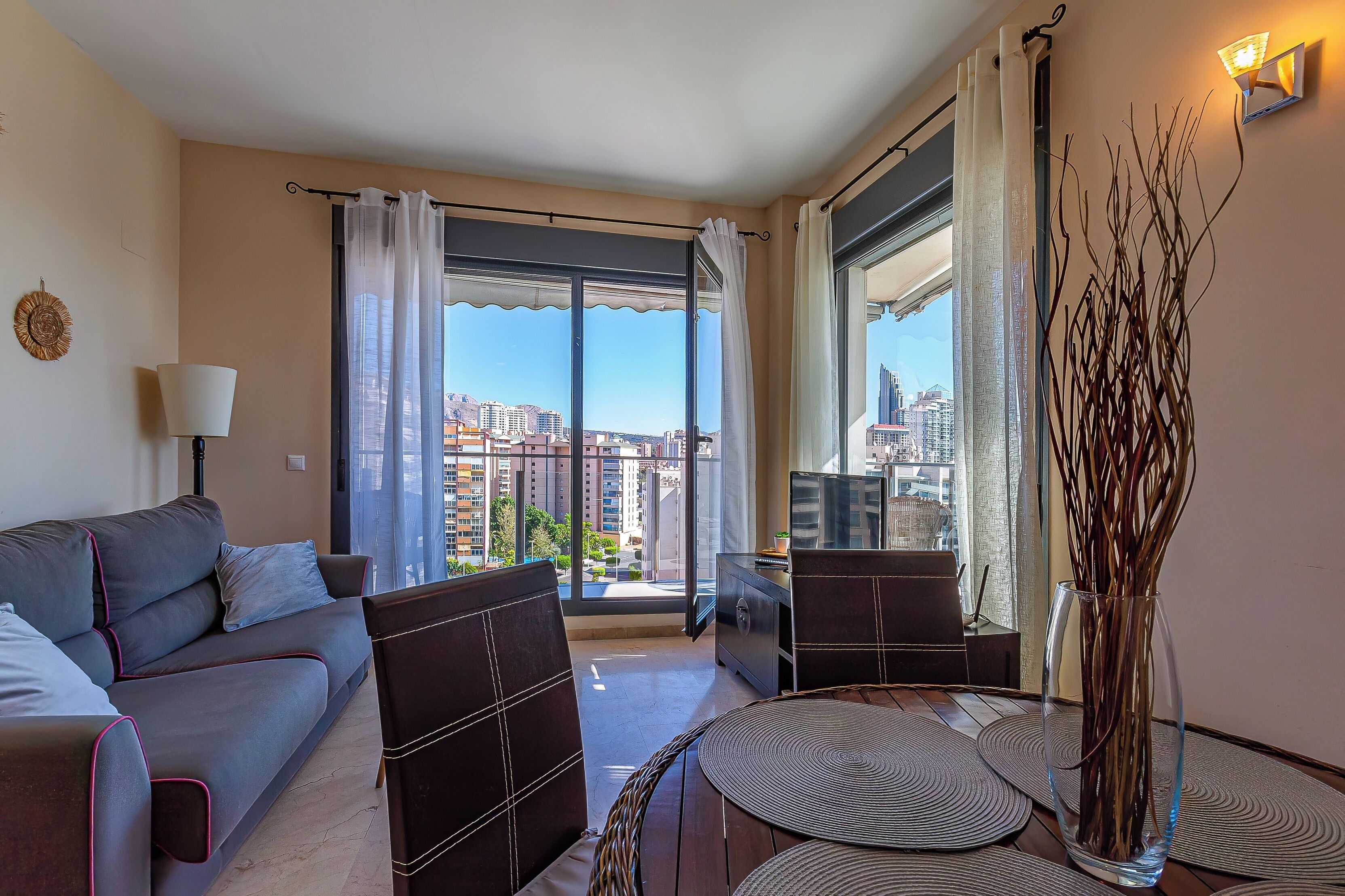 A modern living room with a sofa, chairs, a table, a vase, and a view of a cityscape through large windows.