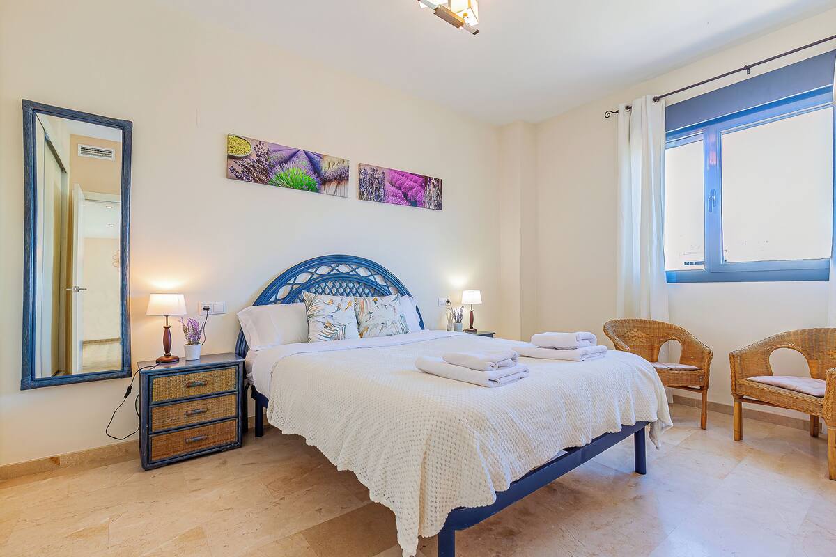 Une chambre à coucher comprenant un lit, des tables de chevet, un miroir et deux fauteuils en osier.