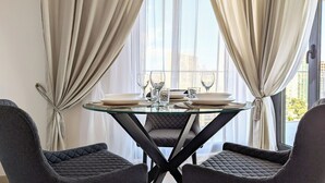 A dining area with a round glass table, two chairs, and a view of buildings through large windows. - Cala Alta Apartments (Villajoyosa)