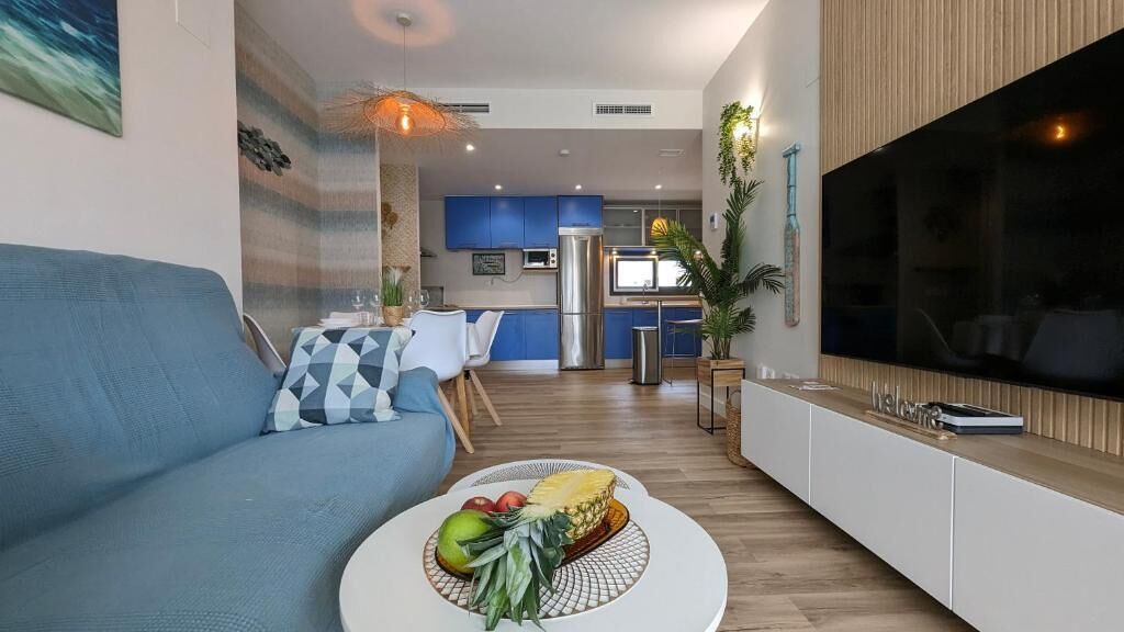 Una sala de estar moderna con un sofá, una mesa de centro con fruta, un televisor y una cocina al fondo.