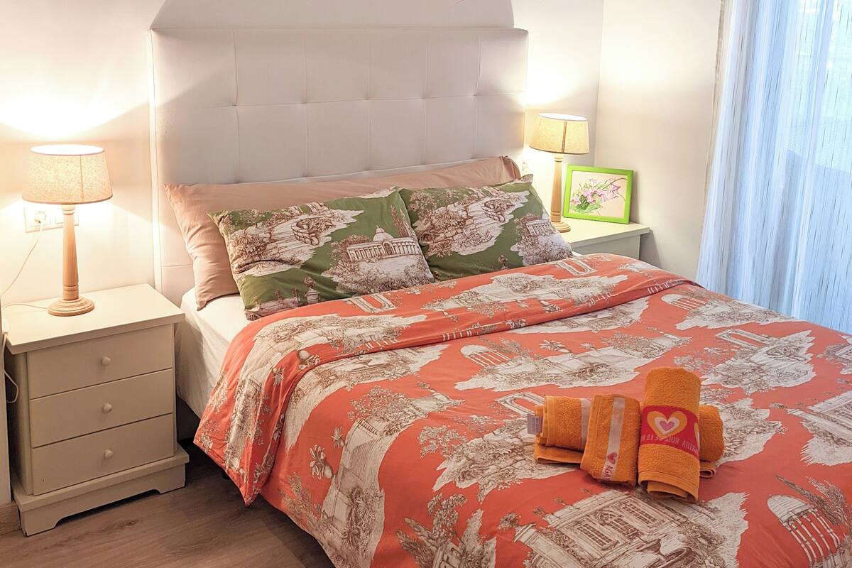 Une chambre à coucher avec un lit, des tables de chevet, une lampe, une fenêtre avec des rideaux et un ventilateur de plafond.